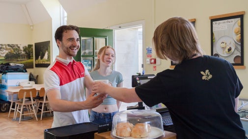 A café counter where a staff member is handing a drink to a customer. On the counter, there is a glass dome with scones inside and a payment terminal. In the background, there are tables, chairs, and posters on the wall. The setting is bright and casual.